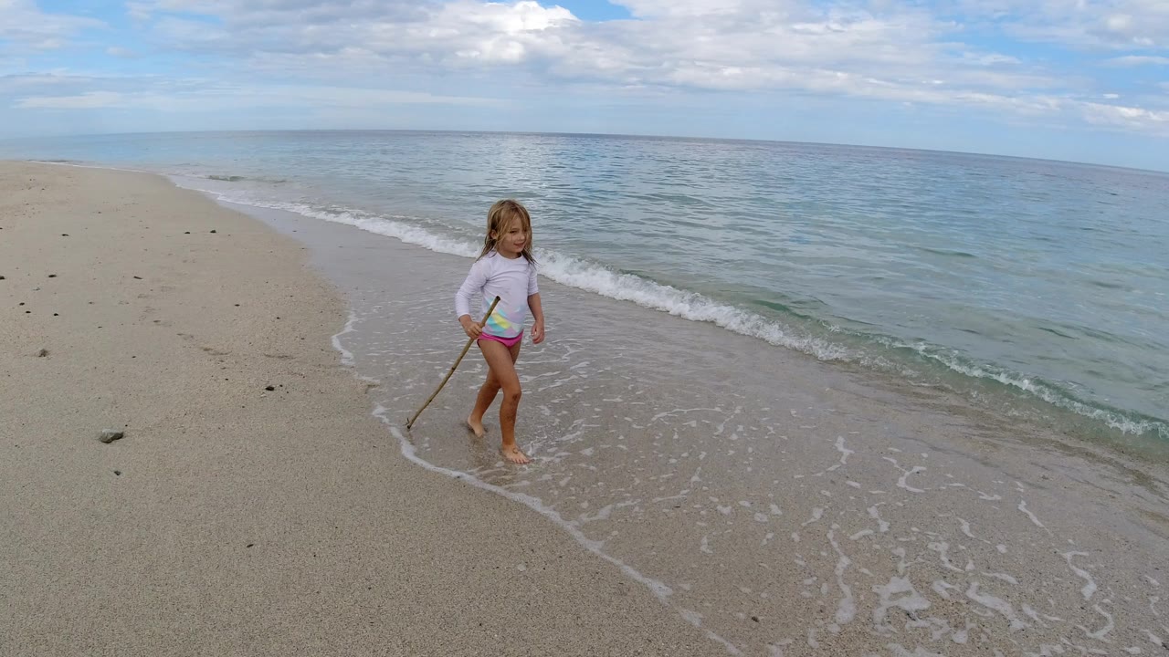A young child in swimwear holds a long stick while standing near small waves on a sandy beach in Los Cabos, Mexico.
