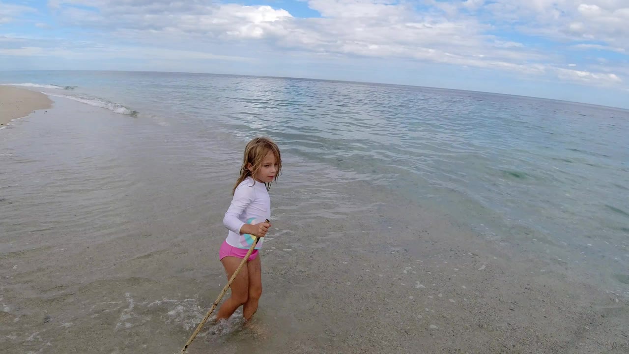 The girl stands closer to the waterline on the Los Cabos beach, holding a stick as thin waves move across pale wet sand.