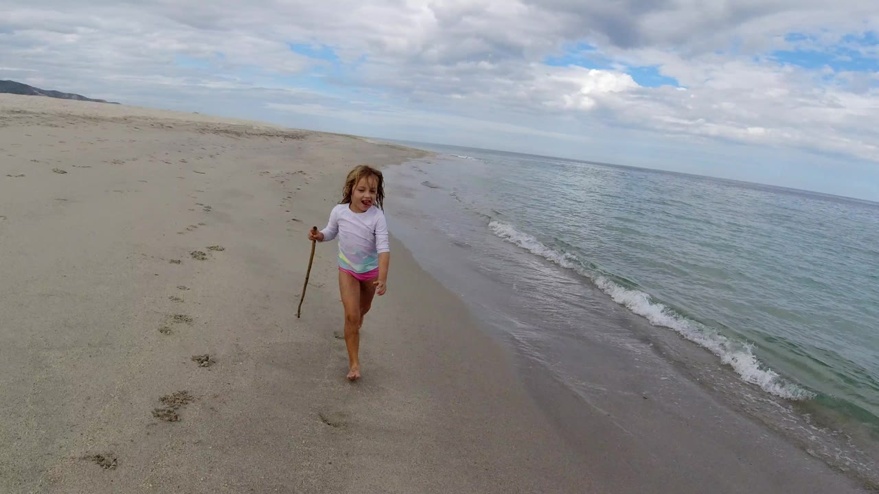 The girl remains near the waterline in Los Cabos, facing the camera with a stick in one hand as gentle surf runs along the right side of frame.