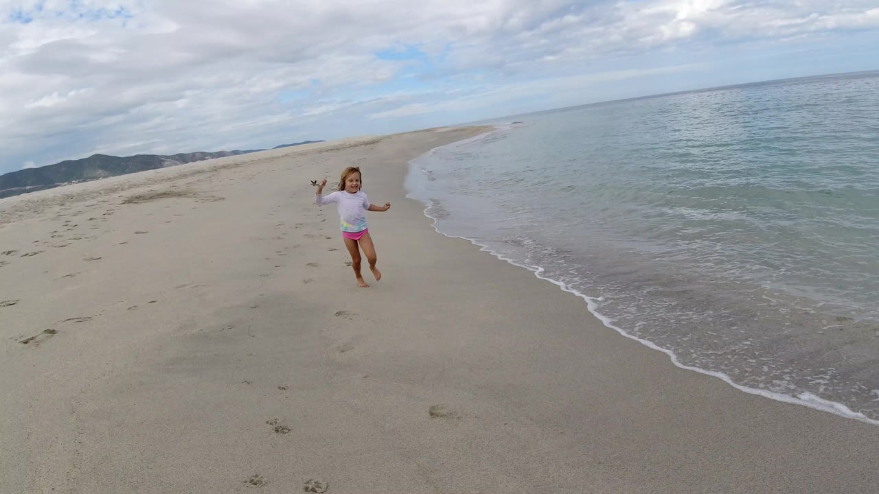 The girl continues along the Los Cabos shoreline, still holding the stick, as a thin line of foam curves up the beach beside her.