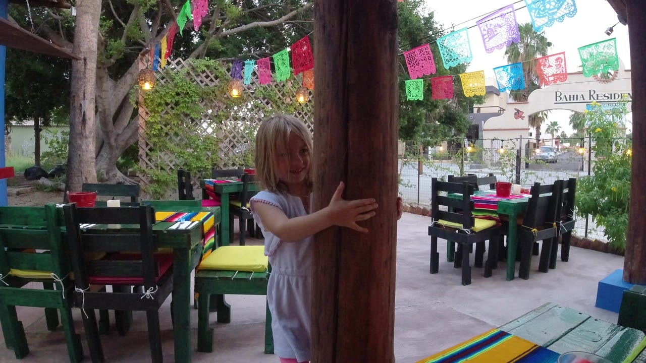 At the start of the Los Cabos clip, a young girl stands partly behind a large wooden post on a restaurant patio with colorful tables and hanging paper banners.