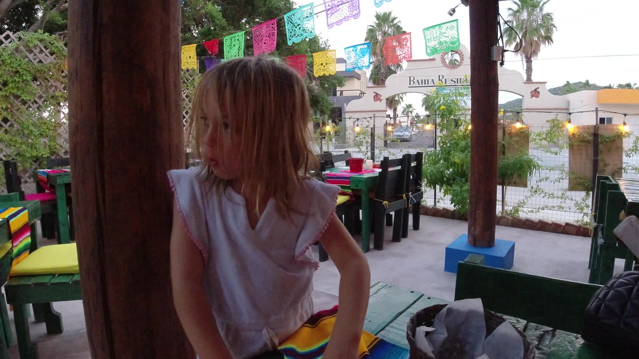 The child remains near a central wood post on the Los Cabos covered deck, looking off to the side while green benches and hanging paper banners sit behind her.