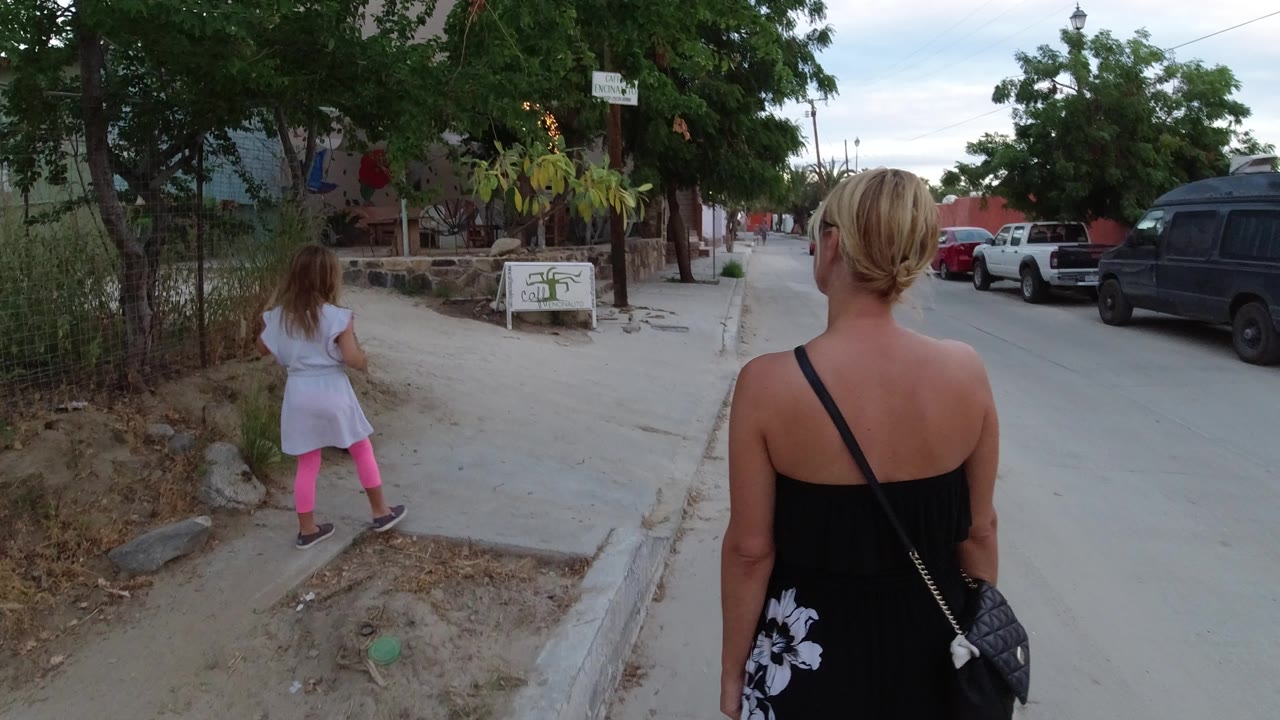 At 0:01 in Los Cabos, Mexico, the child moves along the curb ahead of the woman, with a small roadside sign, trees, and parked cars lining the street.