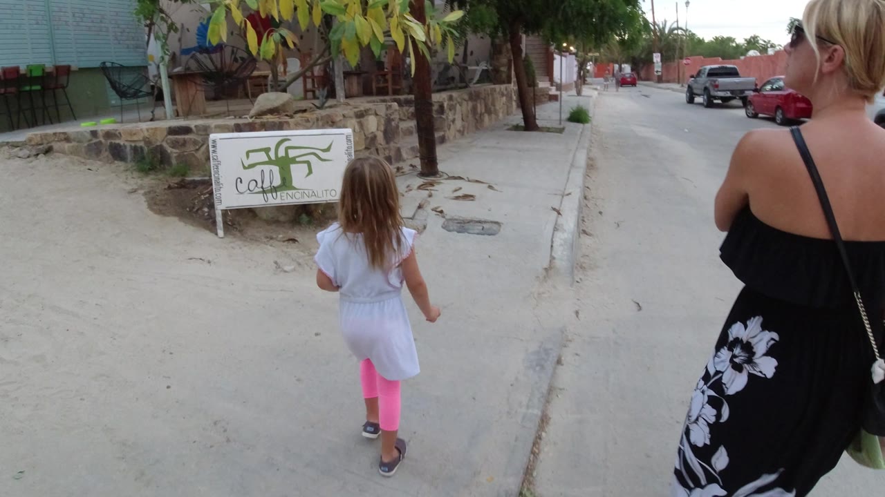 The child continues down the Los Cabos sidewalk with the adult woman partly cropped on the right, passing a low stone wall, leafy trees, a roadside sign, and parked vehicles along the street.
