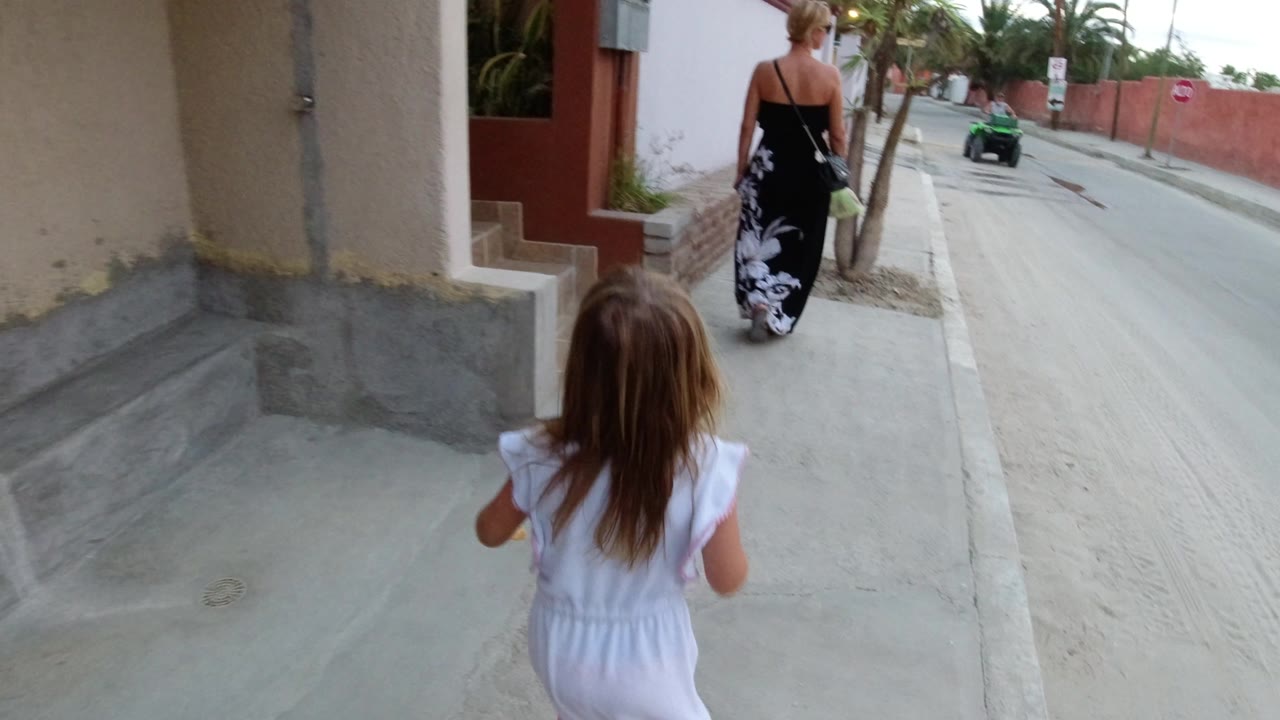 Young girl walking behind a woman on a sidewalk beside a street.