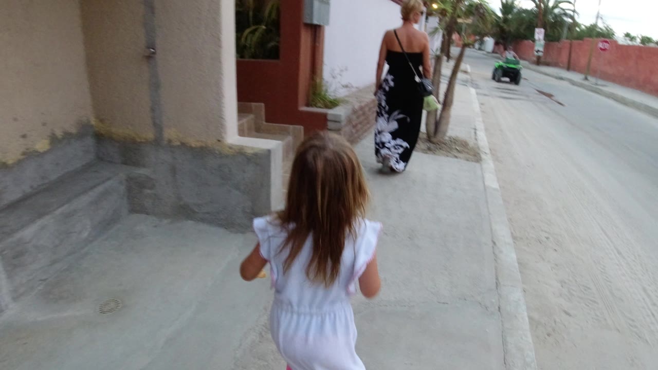 The Los Cabos sidewalk view continues with the child centered in the foreground, the woman ahead, and a green ATV visible farther down the road.