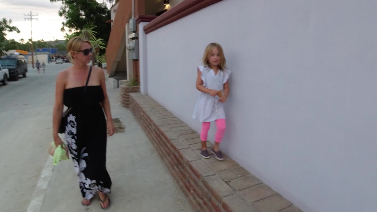 In Los Cabos, Mexico, the woman stands in the left foreground while the young girl remains near the white wall in the midground, with cars and a street behind them.