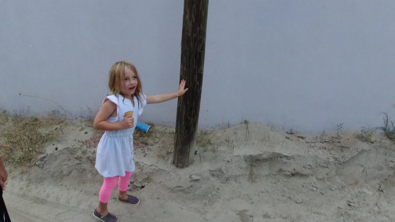 A young girl in a white dress and pink leggings stands beside a wood utility pole on sandy ground in Los Cabos, Mexico, with a pale wall behind her.