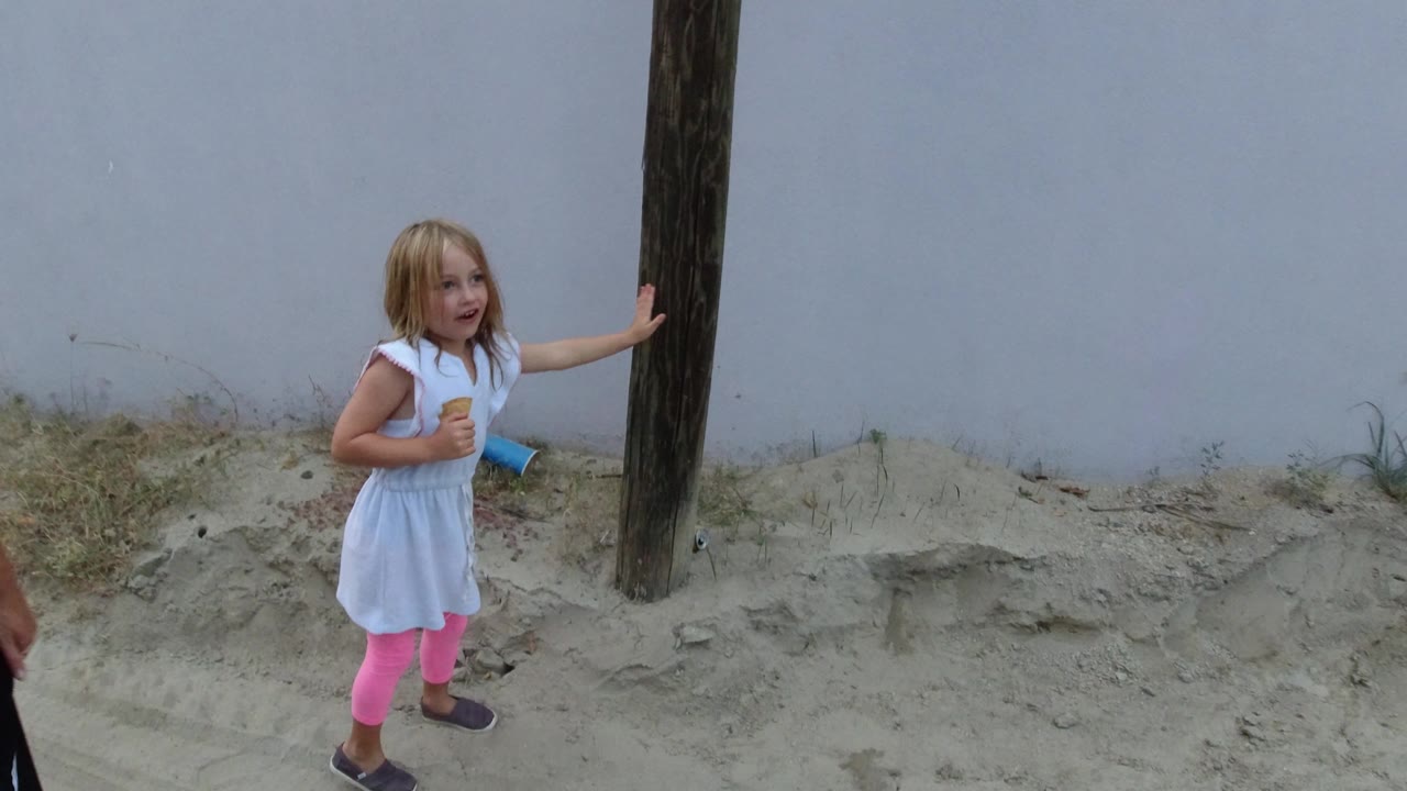 The girl holds one hand against the wood pole while standing on the sandy roadside in Los Cabos, Mexico, with the pale wall filling the background.