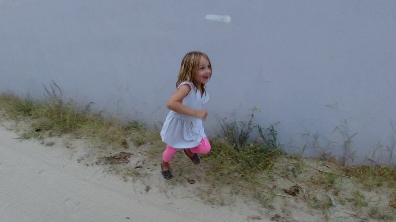 Young girl walking beside a white wall on a sandy street.