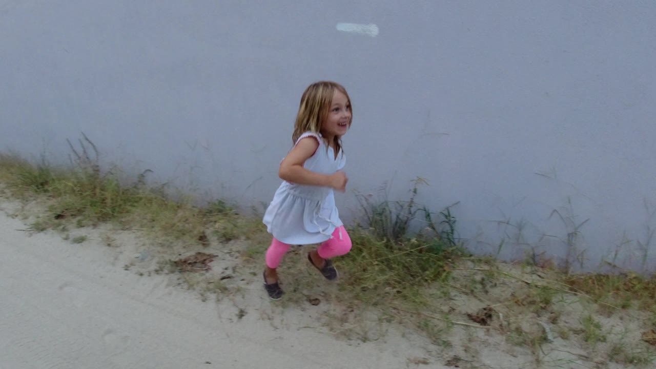 The girl continues forward near the white wall, with sparse grass and pale sand visible under soft daylight in Los Cabos, Mexico.