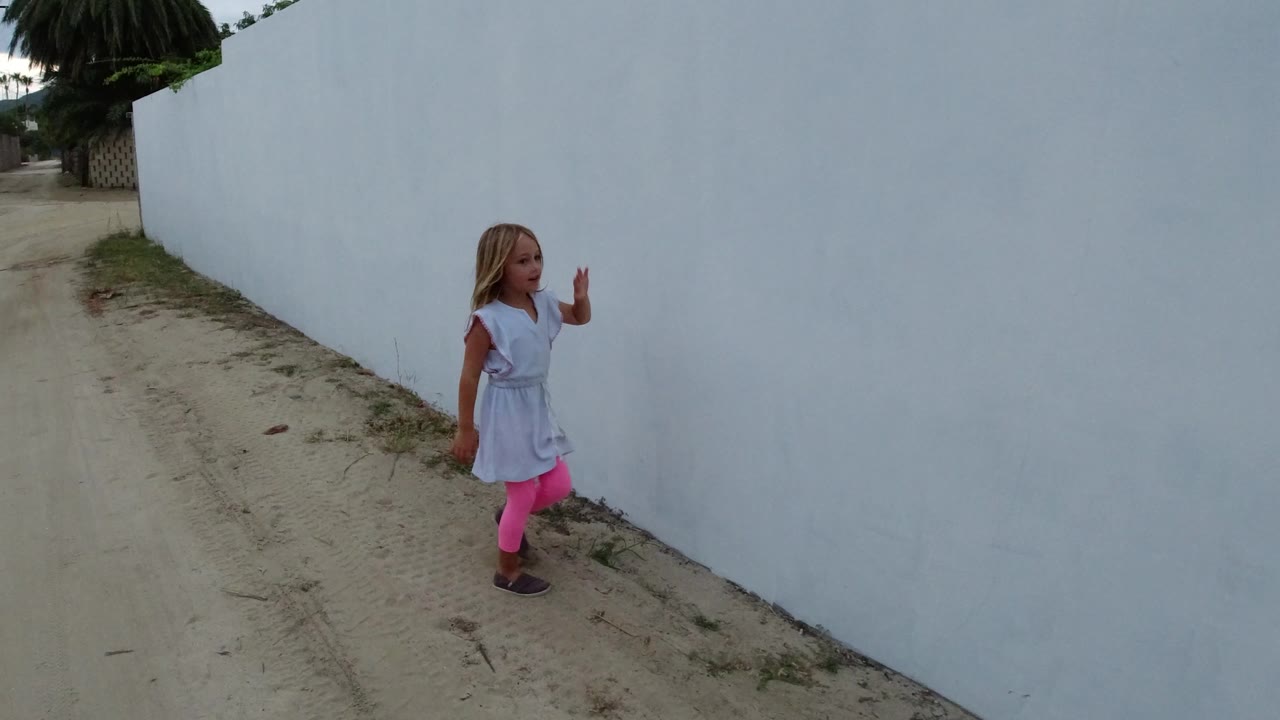 Young child beside a tall white wall on a sandy road.