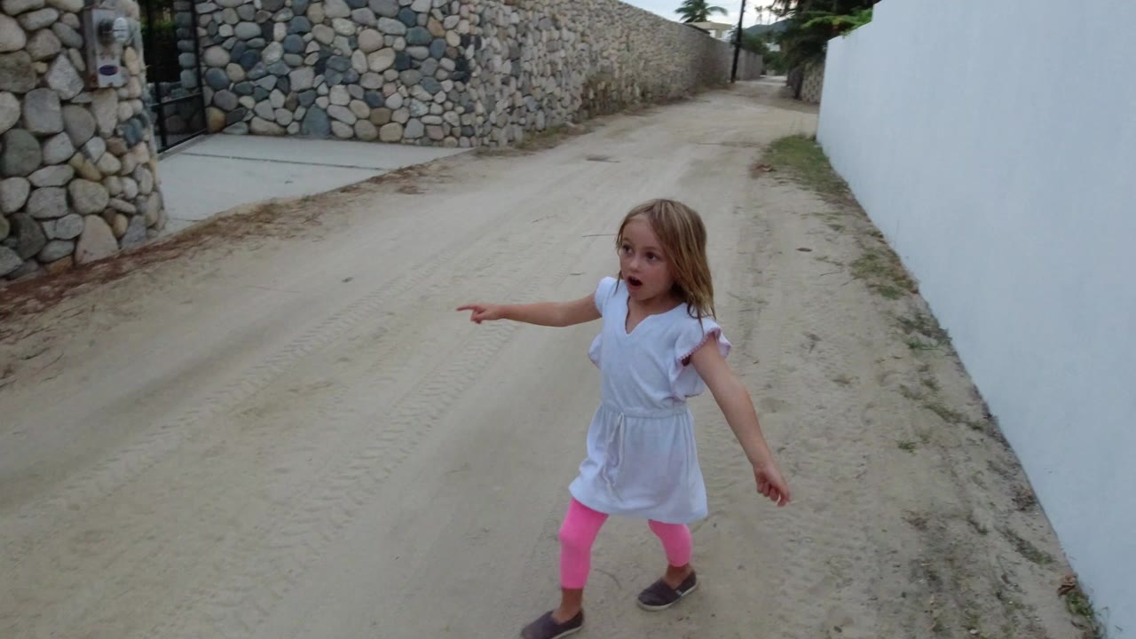 In Los Cabos, a young blonde girl in a white dress and pink leggings points across a sandy lane bordered by stone and white walls.