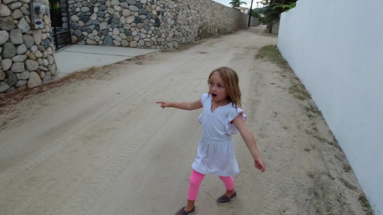 The Los Cabos lane remains in view as the young girl continues pointing, with a stone boundary wall on one side and a plain white wall on the other.