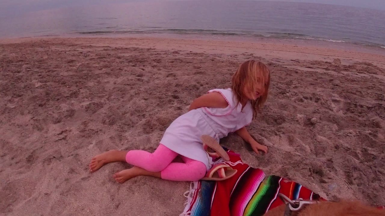 Young girl in pink leggings sits on the sand beside a colorful striped blanket on a Los Cabos beach, with low waves and muted afternoon light behind her.