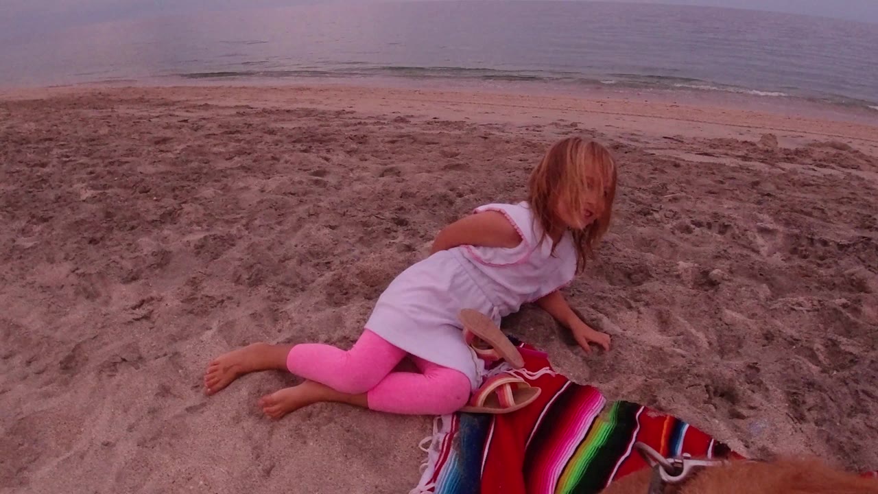 The girl remains seated on the Los Cabos sand near the striped blanket, turning slightly toward the camera while the shoreline runs across the background.