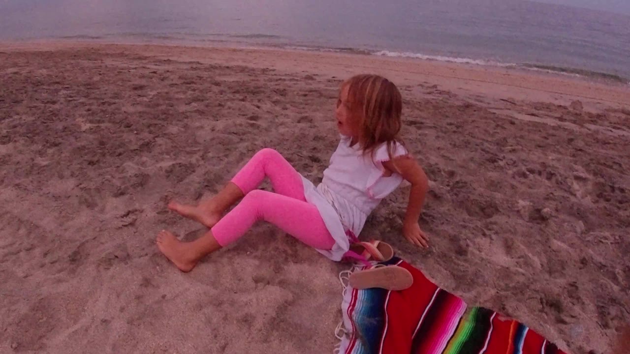 The child remains seated on uneven sand in Los Cabos, turned slightly toward the water with a bright striped blanket and sandal at the lower edge.