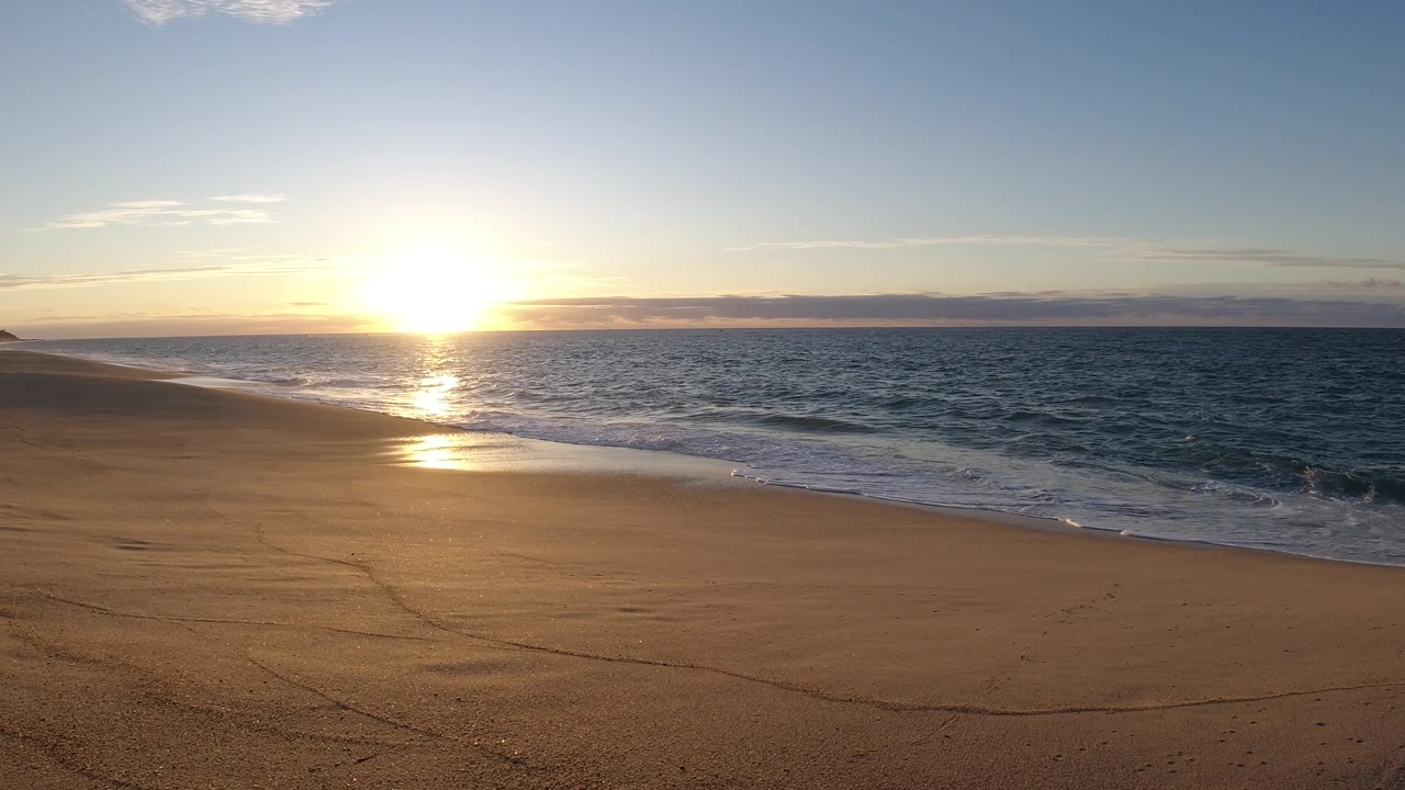 In Los Cabos, Mexico, the sun reflection brightens the wet sand while a thin wave line curls onto the empty beach with no visible people.