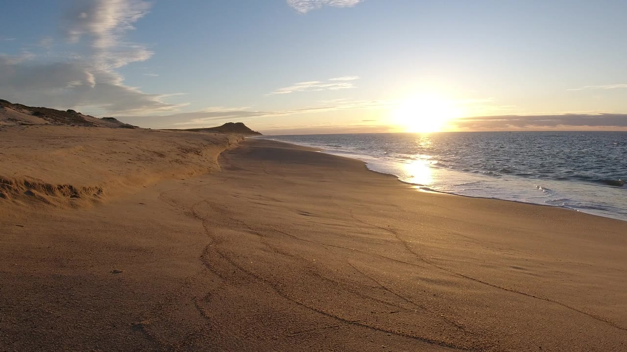 Wide view of a sandy Los Cabos beach with the low sun reflecting across the ocean and wet shoreline, with no visible people.