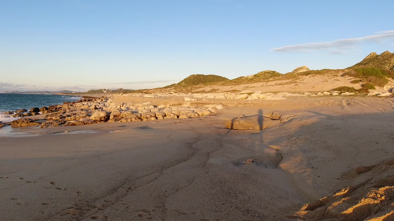 Wide Los Cabos beach view at the start of the clip, with sand channels in front, rocky shoreline and ocean to the left, sunlit hills behind, and no visible people.