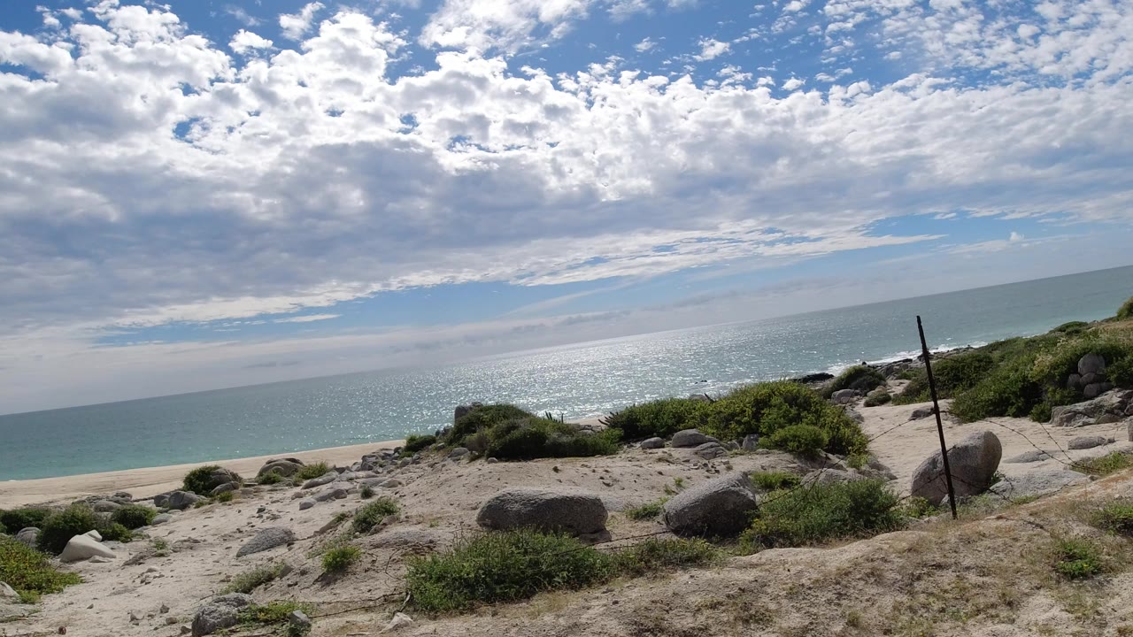 Los Cabos sandy beach overlook with scattered boulders, low shrubs, a thin fence post, bright ocean glare, layered clouds, and no visible people.