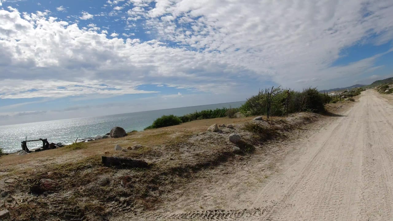 The Los Cabos coastal road continues ahead with the ocean visible to the left, scattered rocks and scrub along the shoulder, pale tire-marked dirt, and no visible people.