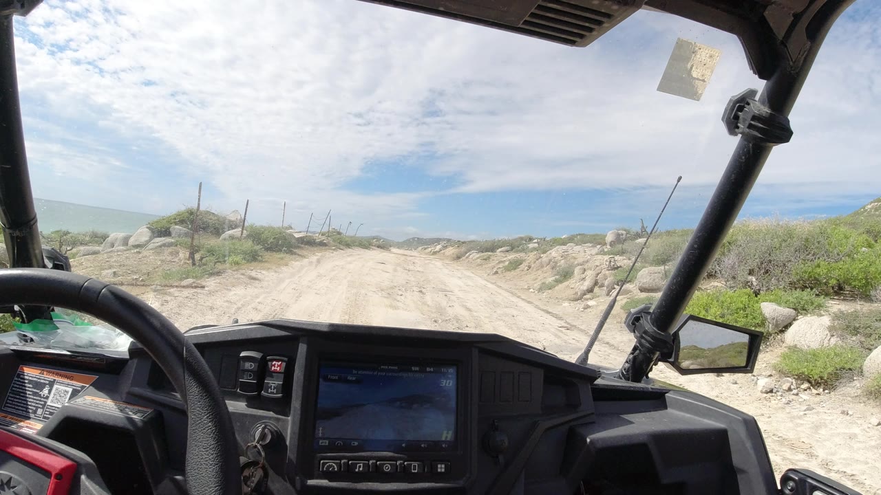 Los Cabos dirt road seen from a vehicle interior, with black dashboard controls, roll cage bars, low shrubs, rocky roadside, pale ocean line, and no visible people.