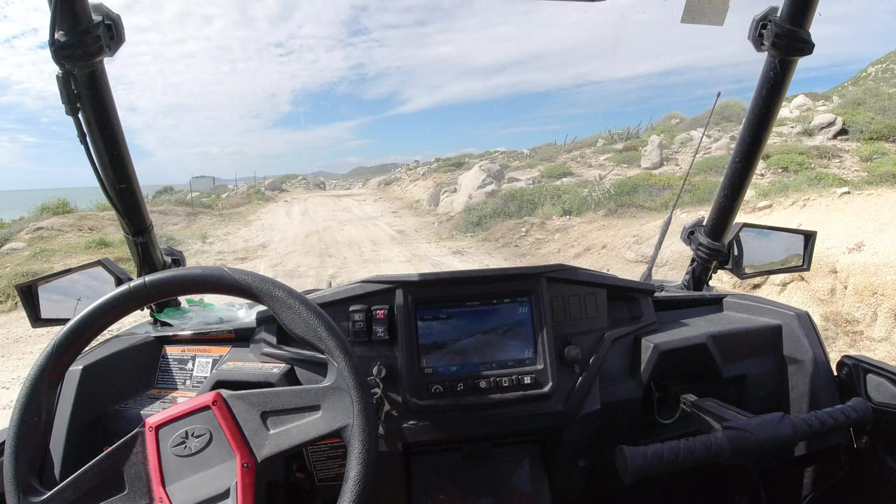 A forward cabin view in Los Cabos, Mexico shows the dashboard screen and steering wheel in the foreground, with a pale sandy track bending through rocky scrub and no visible people.