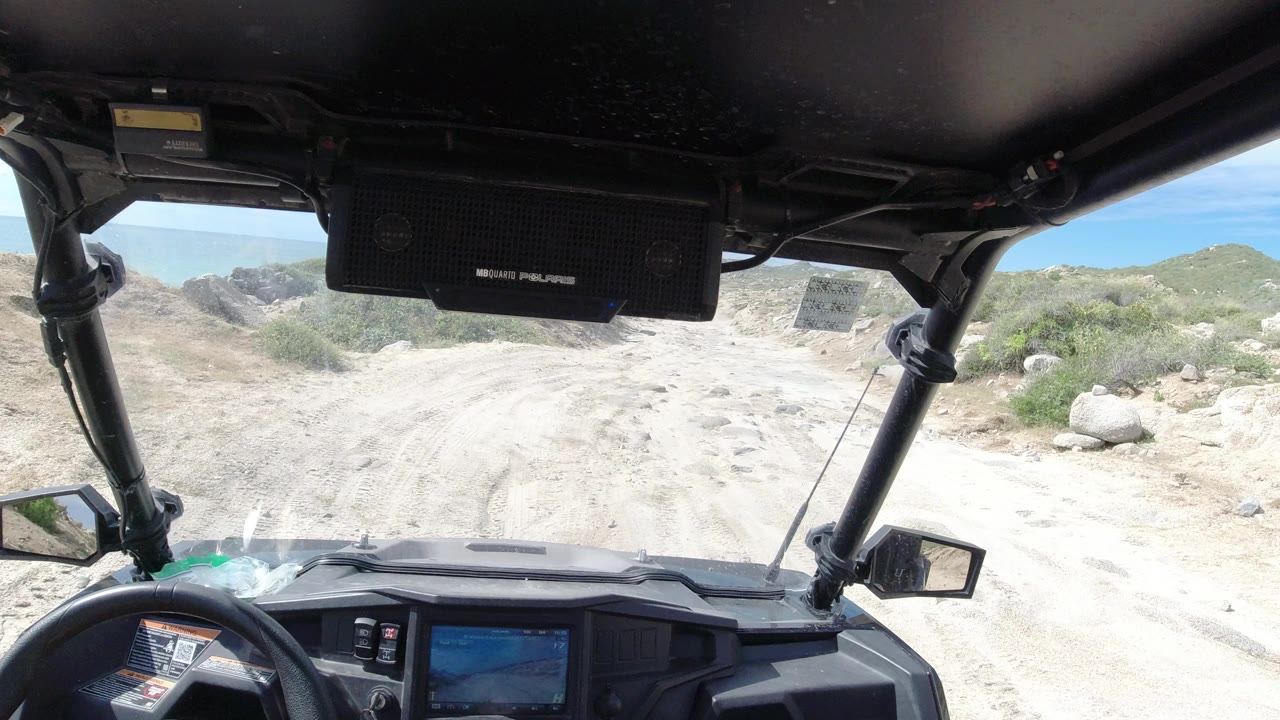 Dashboard and roll-cage view along a sunlit Los Cabos dirt track, with scrubby desert plants, rocks, low hills, and no visible people.