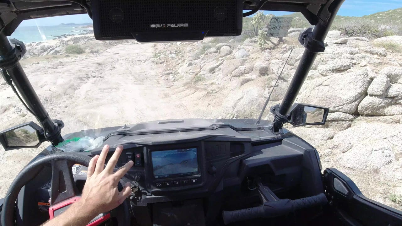 A hand reaches toward the dashboard while the vehicle faces a rough sandy trail in Los Cabos, Mexico, with rocks and beach light beyond.