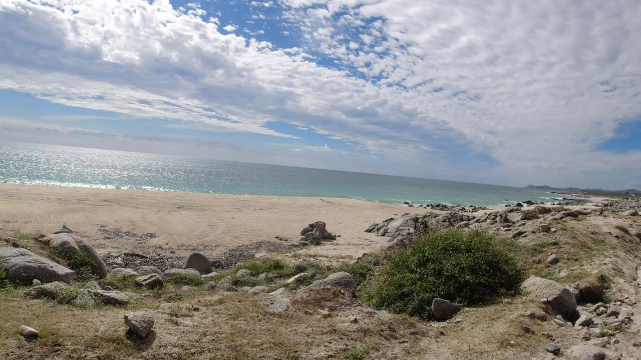 A Los Cabos, Mexico cliffside view looks over pale beach and turquoise ocean, with rocks and scrub in the foreground and no visible people.