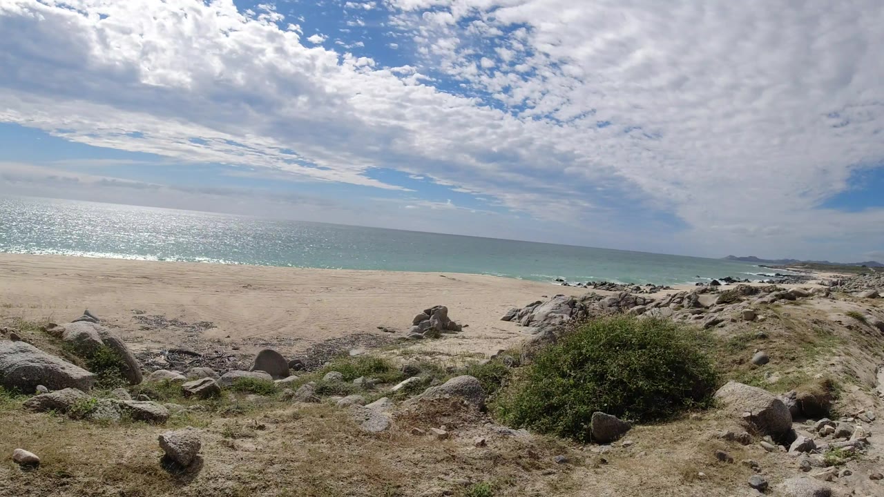 The Los Cabos shoreline remains wide in frame, with foreground boulders, dry cliff soil, low brush, bright sea glare, and no visible people.
