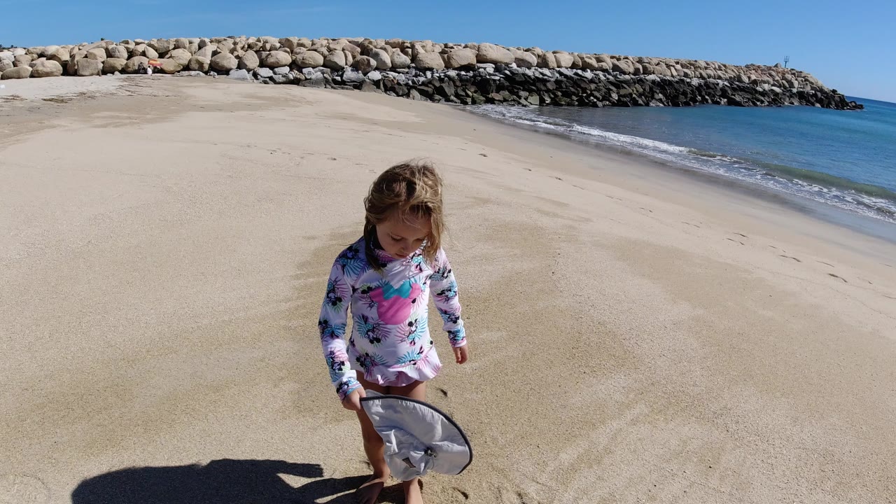 A young girl stands on a Los Cabos beach beside a floppy hat and shoes, with pale sand, blue water, and a rock breakwater in the background.