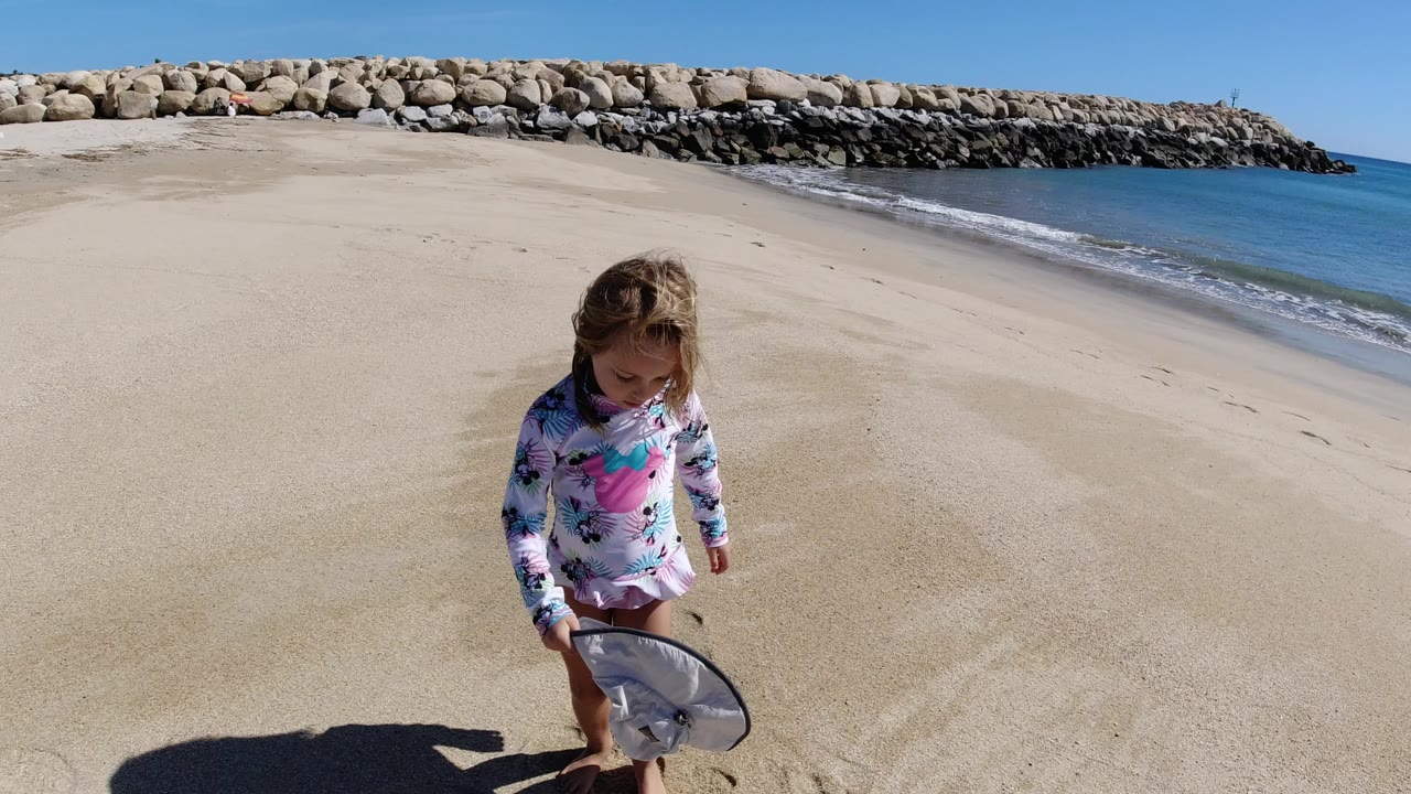The girl looks down toward a hat and shoes on the sand while the Los Cabos shoreline and rock breakwater remain behind her.