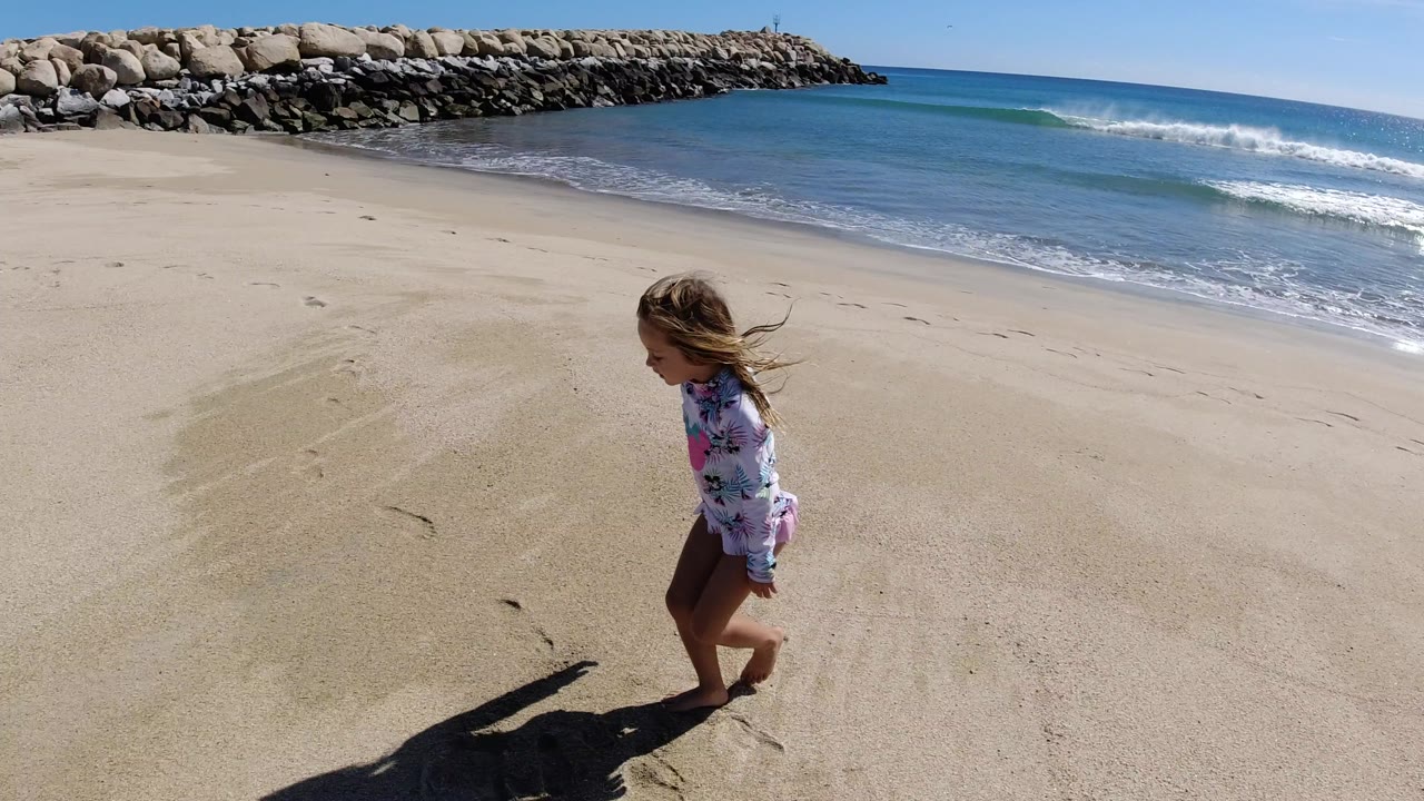 The girl continues along the Los Cabos shoreline, positioned lower in frame with pale sand in front, surf at midground, and the rock breakwater across the top.