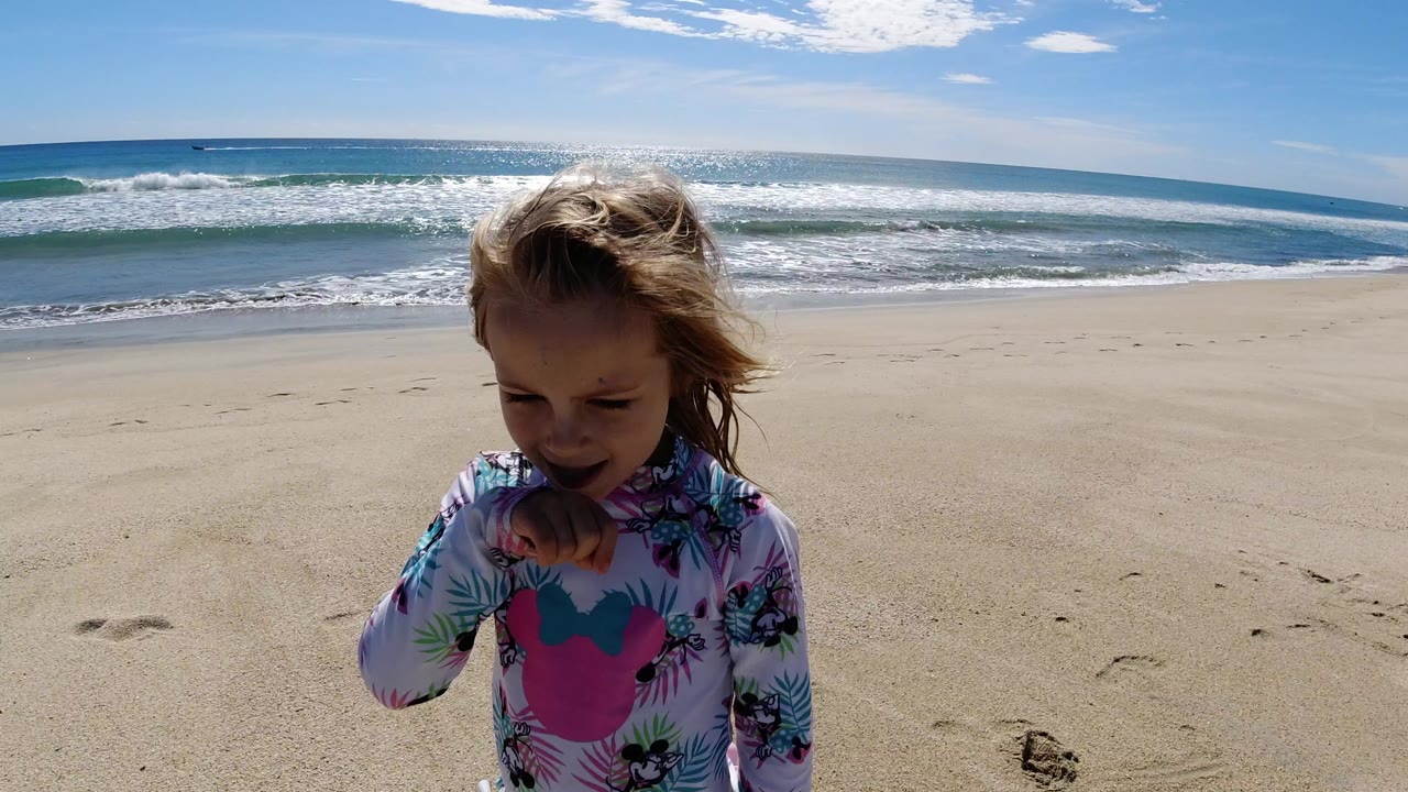 Young girl in a patterned swim shirt stands on sand with surf behind her.