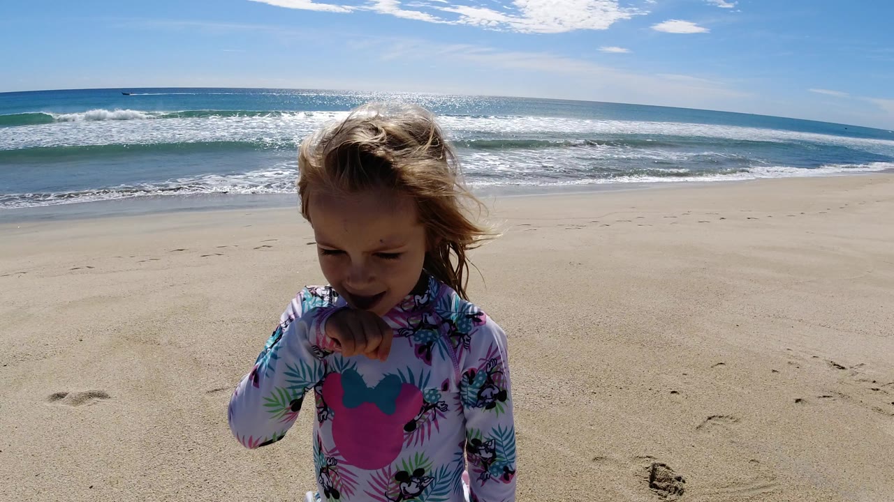 At 0:01 in Los Cabos, Mexico, the girl is centered closer to camera with her hand near her mouth, standing on pale sand before blue water and a low horizon.