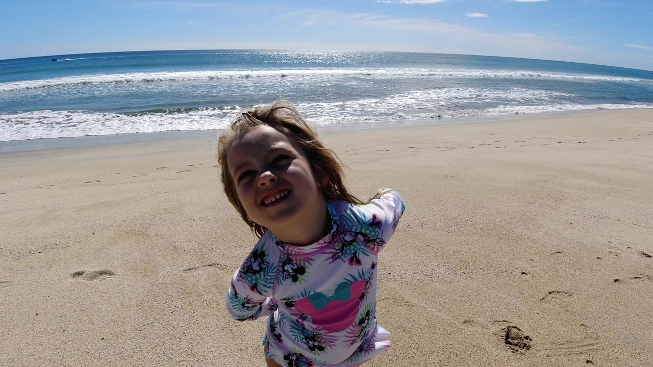 A young girl in a floral swim shirt stands close to the camera on a Los Cabos beach, with pale sand, breaking waves, and blue sky behind her.