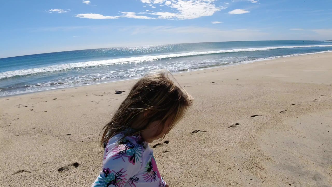 A young girl in a patterned swim top walks near the lower left of a Los Cabos beach frame, with bright ocean surf, pale sand, and a wide blue sky behind her.