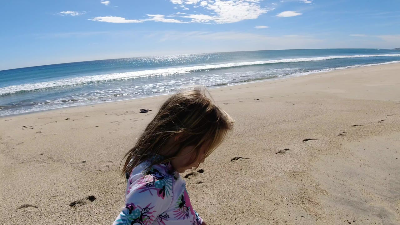 The girl remains low in the frame on the Los Cabos sand while a line of white surf advances across the background and footprints mark the beach.