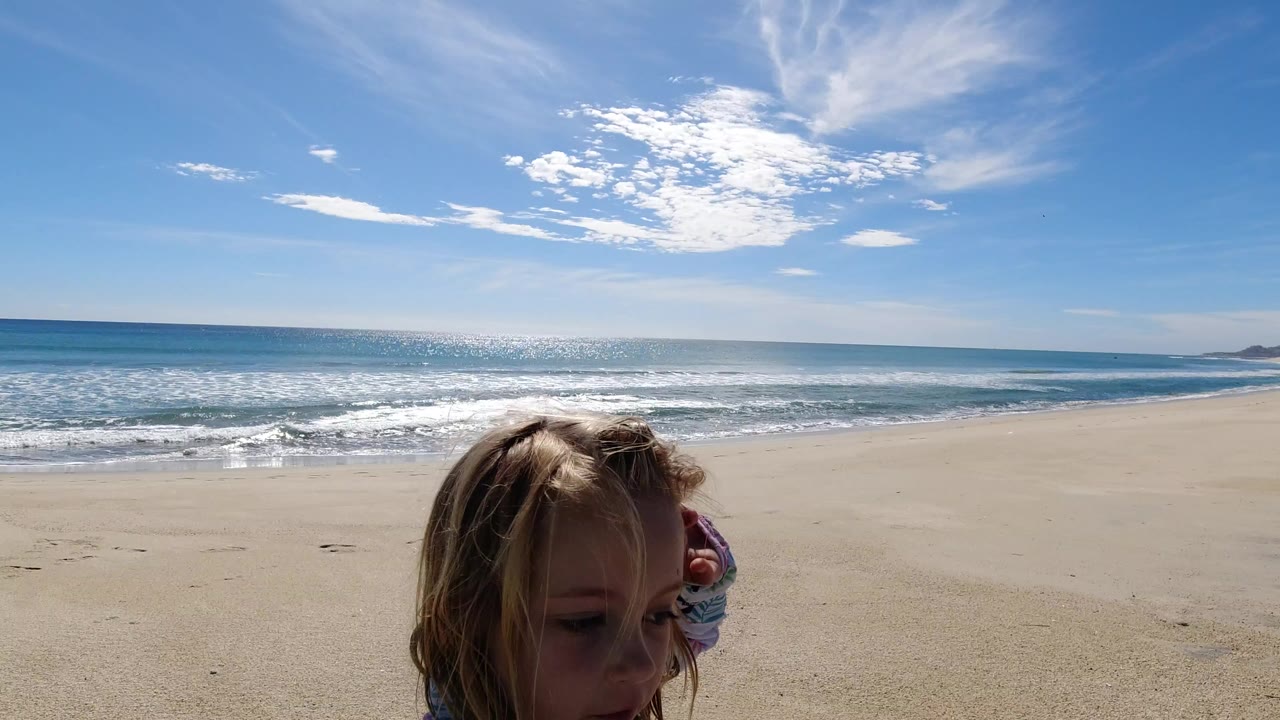 At the start of the Los Cabos beach clip, a young girl appears close in the foreground with ocean surf, pale sand, and bright clouded sky behind her.