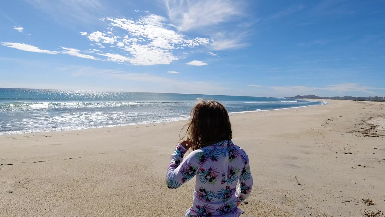 The girl remains centered from behind on a Los Cabos beach, facing the long curve of shoreline as sunlight reflects across the ocean.