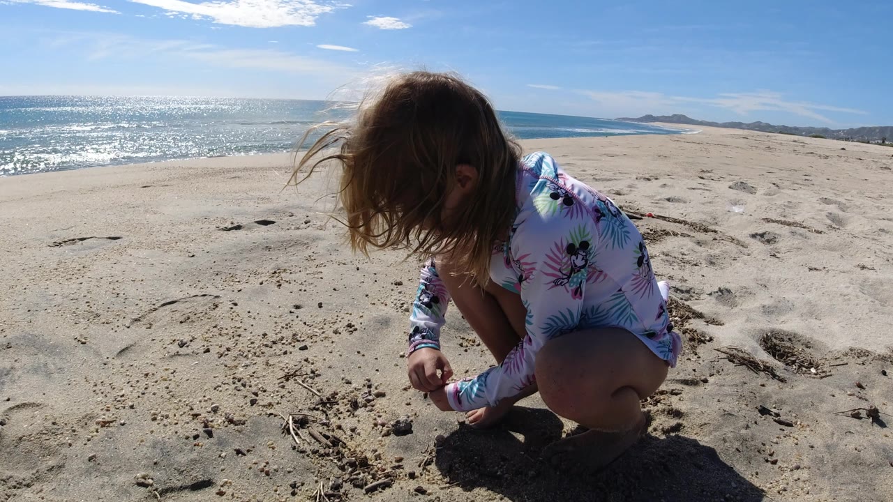 The girl remains crouched low on the Los Cabos shoreline, looking down as one hand works through the sand and sun glints across the water behind her.