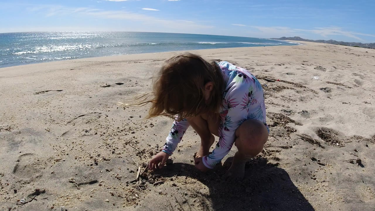 Young girl crouching in sand beside a bright ocean shoreline.