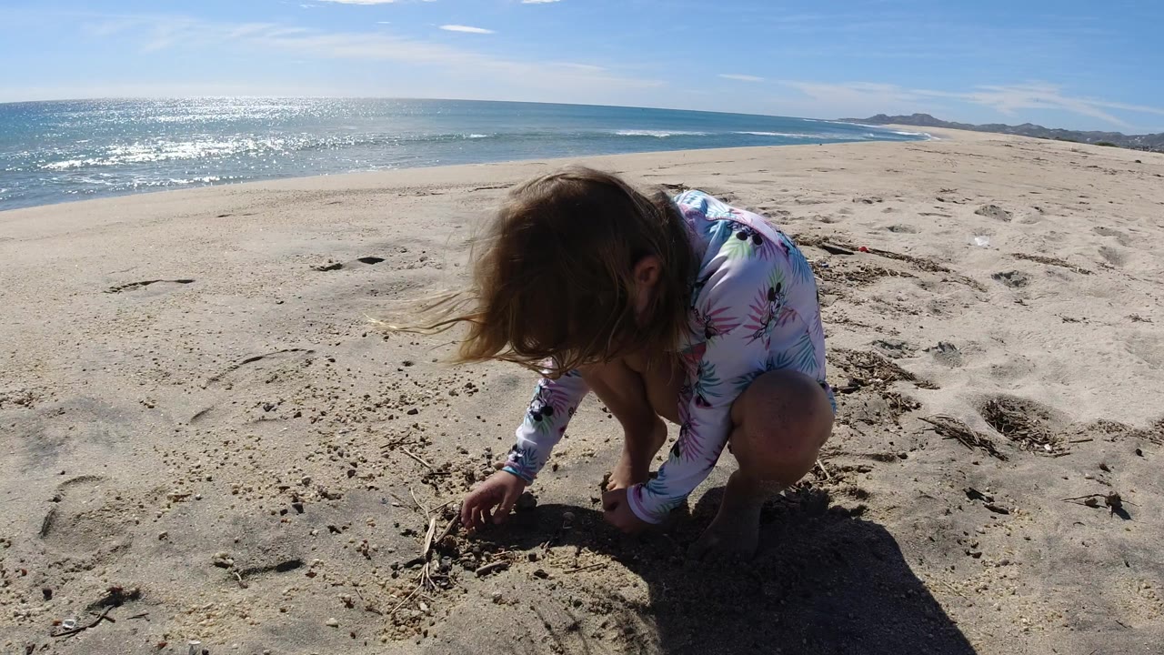 The girl leans lower toward the sand in Los Cabos, with her hair falling forward and the glittering ocean horizon stretching across the background.