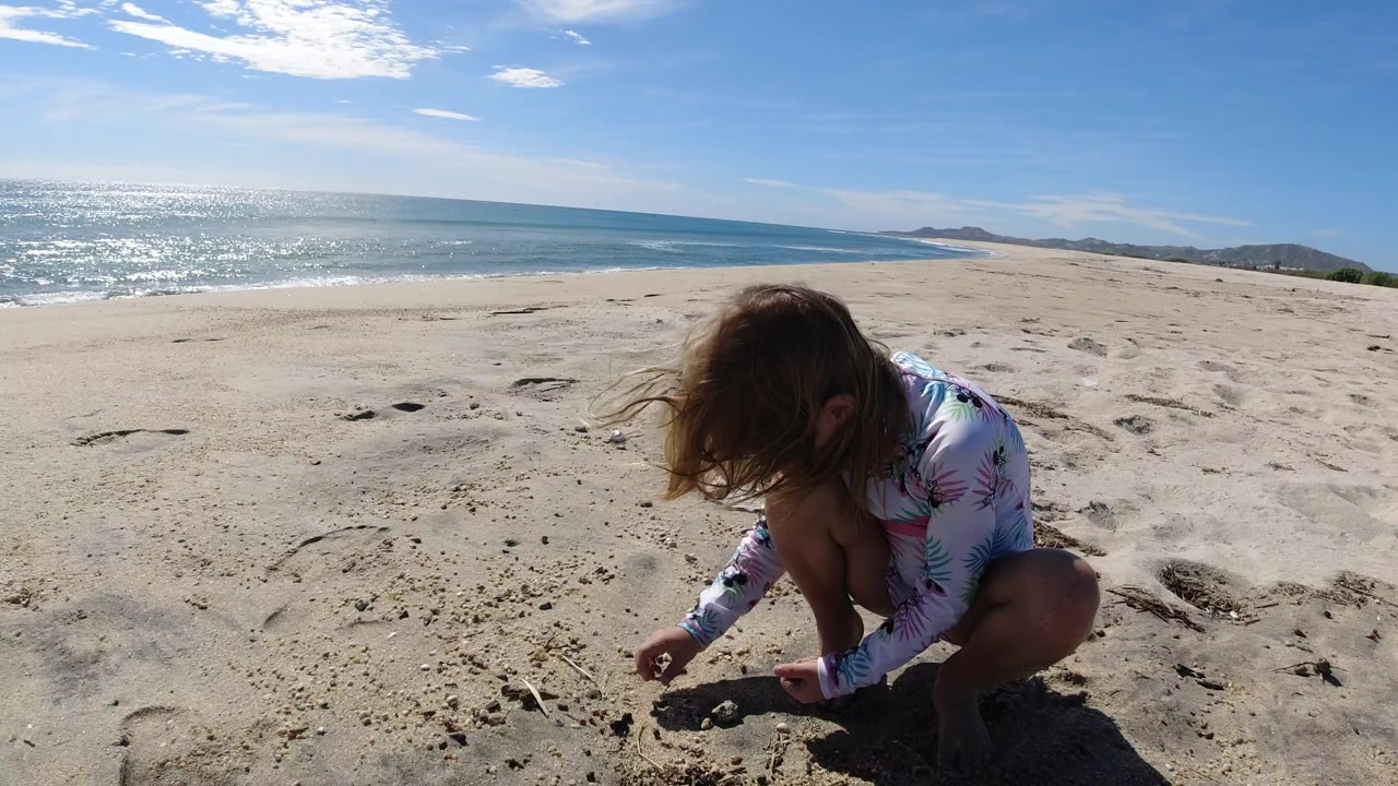 A blonde child crouches low in the sand on a Los Cabos beach, with bright ocean water, pale sand, and distant hills behind them.