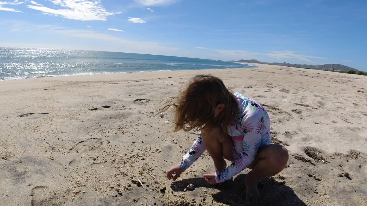 The child remains crouched near small sand marks while the Los Cabos shoreline and bright blue sea run across the background.