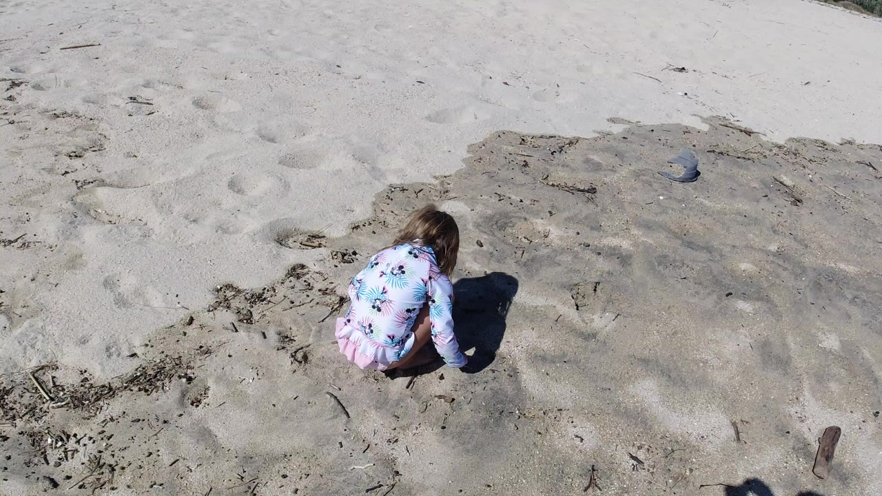 Child crouching on pale beach sand near scattered seaweed and footprints.