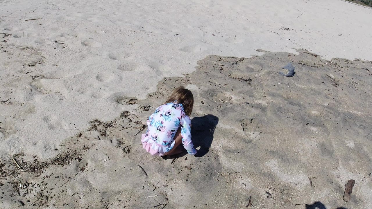 The child remains crouched over the Los Cabos beach sand, with their floral swimsuit bright against muted sand and scattered organic debris.