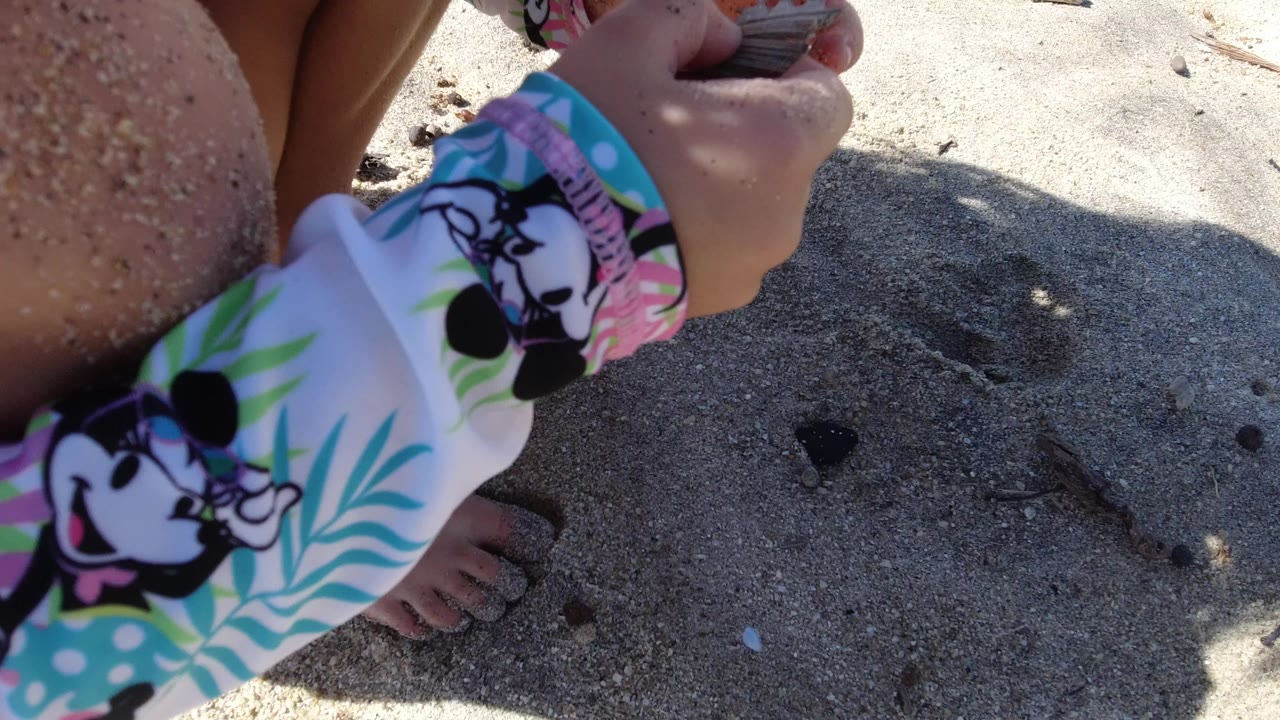 A close beach frame shows the child crouched in Los Cabos, Mexico, with a colorful sleeve, sandy knee, bare foot, and hand near small debris on the sand.